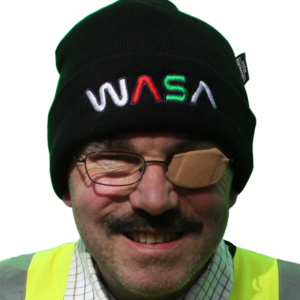 A man in hi-viz jacket wearing a black hat with the letter W, A, S, A embroidered on it. He is grinning and has sticking plaster over his spectacles for his left eye.