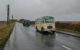 A vintage green and cream coloured bus travels on a wet road followed by a green lorry. on the left hand side of the road are a run of 8 armed telegraph poles.