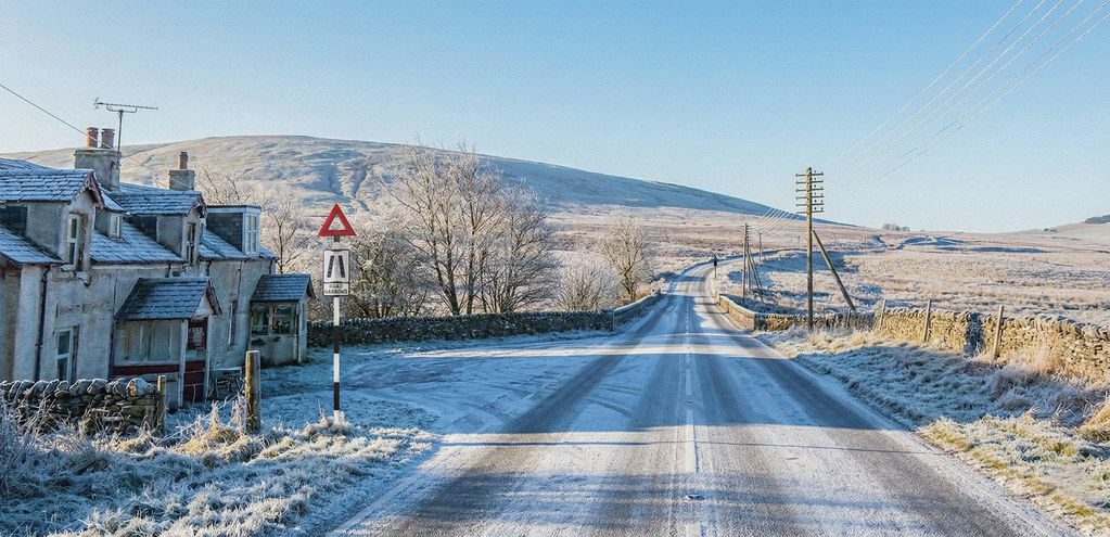 A lightly snowed road heads off into the hills of southern scotland. A dilapidated cottage is on the left and a run of 6 armed telegraph poles on the right. It is a sunny day.