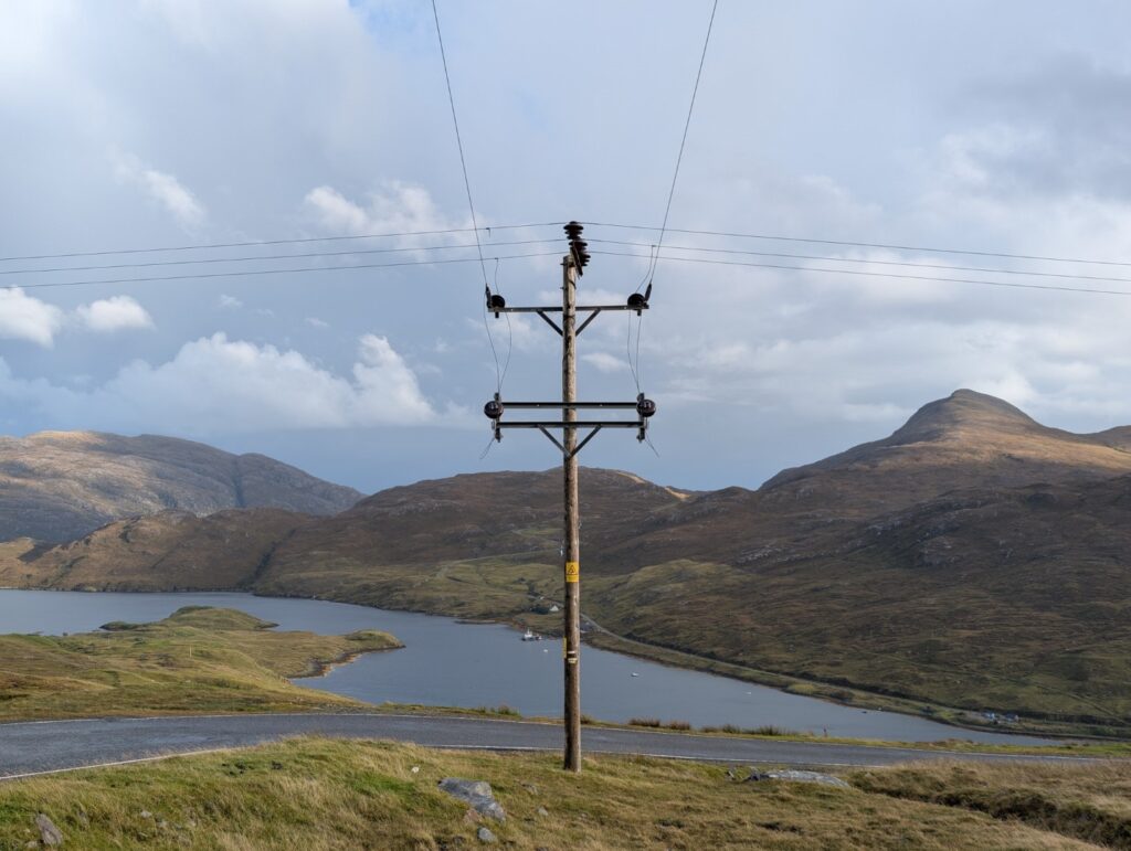 A power pole intersection high above a loch in the highlands of Lewis, Scotland with mountains in the background. Photo by Matthew Pilkington
